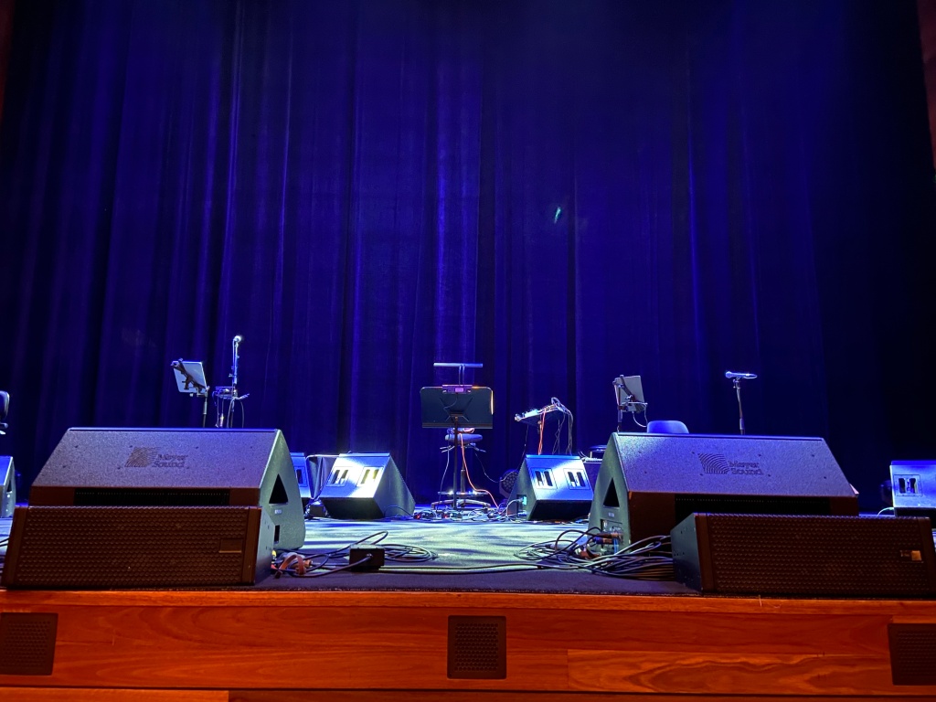 A blue lit stage with a scattering of speakers, music stands, and microphones.